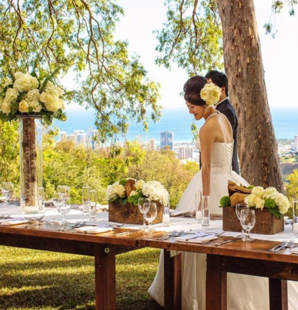 A bride and groom stand at a table, smiling, with a beautiful ocean view in the background.