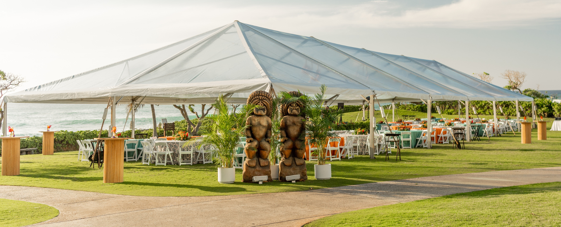 A large tent with tiki decorations, tables, and chairs arranged on the grass for an outdoor event.