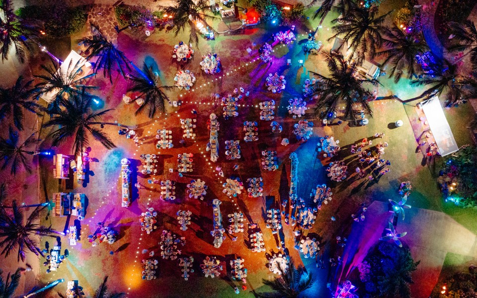 Aerial view of a lively party surrounded by palm trees, showcasing vibrant decorations and people enjoying the celebration.